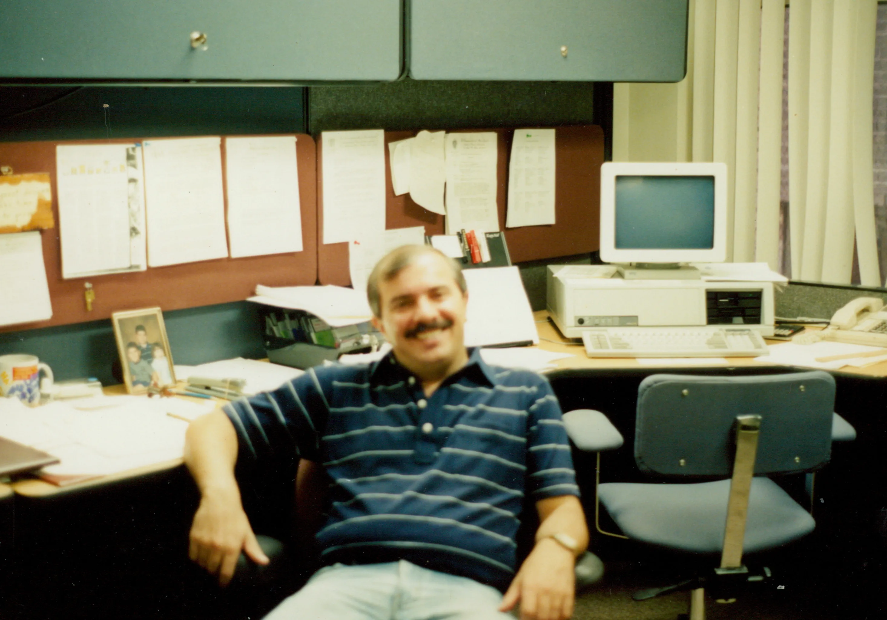 Bert at his office desk, smiling, with a family photo and bulletin board behind him