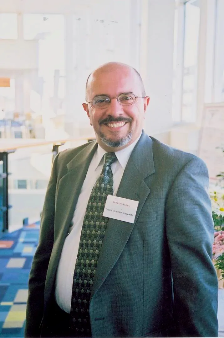 Bert at a professional conference, wearing a green suit with a name badge reading 'Bert Lourenco / Office of Human Resources'